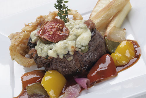 Filet mignon topped with bleu cheese crumbles, served with roasted peppers and yuca fries on a white plate.